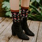 A woman's legs above black booties on a forest path, modeling Gems Of The Forest Bamboo socks in Black with red mushroom and floral details. [lf]