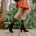 A pair of black heeled booties and Gems Of The Forest Bamboo socks in Black on a wood deck, the pattern of mushrooms and daisies clearly visible at the cuff. [lf]