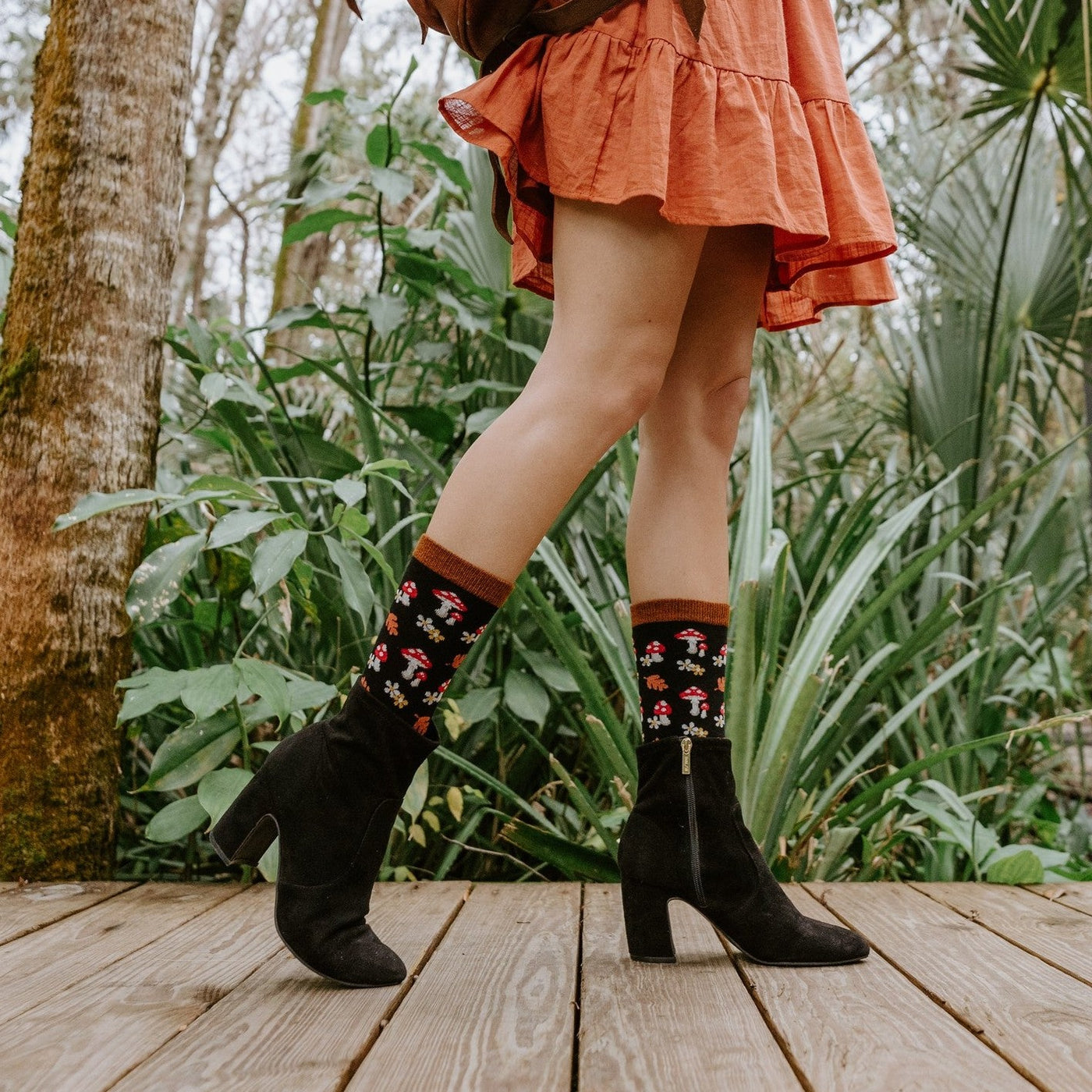 A pair of black heeled booties and Gems Of The Forest Bamboo socks in Black on a wood deck, the pattern of mushrooms and daisies clearly visible at the cuff. [lf]