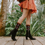 A pair of black heeled booties and Gems Of The Forest Bamboo socks in Black on a wood deck, the pattern of mushrooms and daisies clearly visible at the cuff. [lf]
