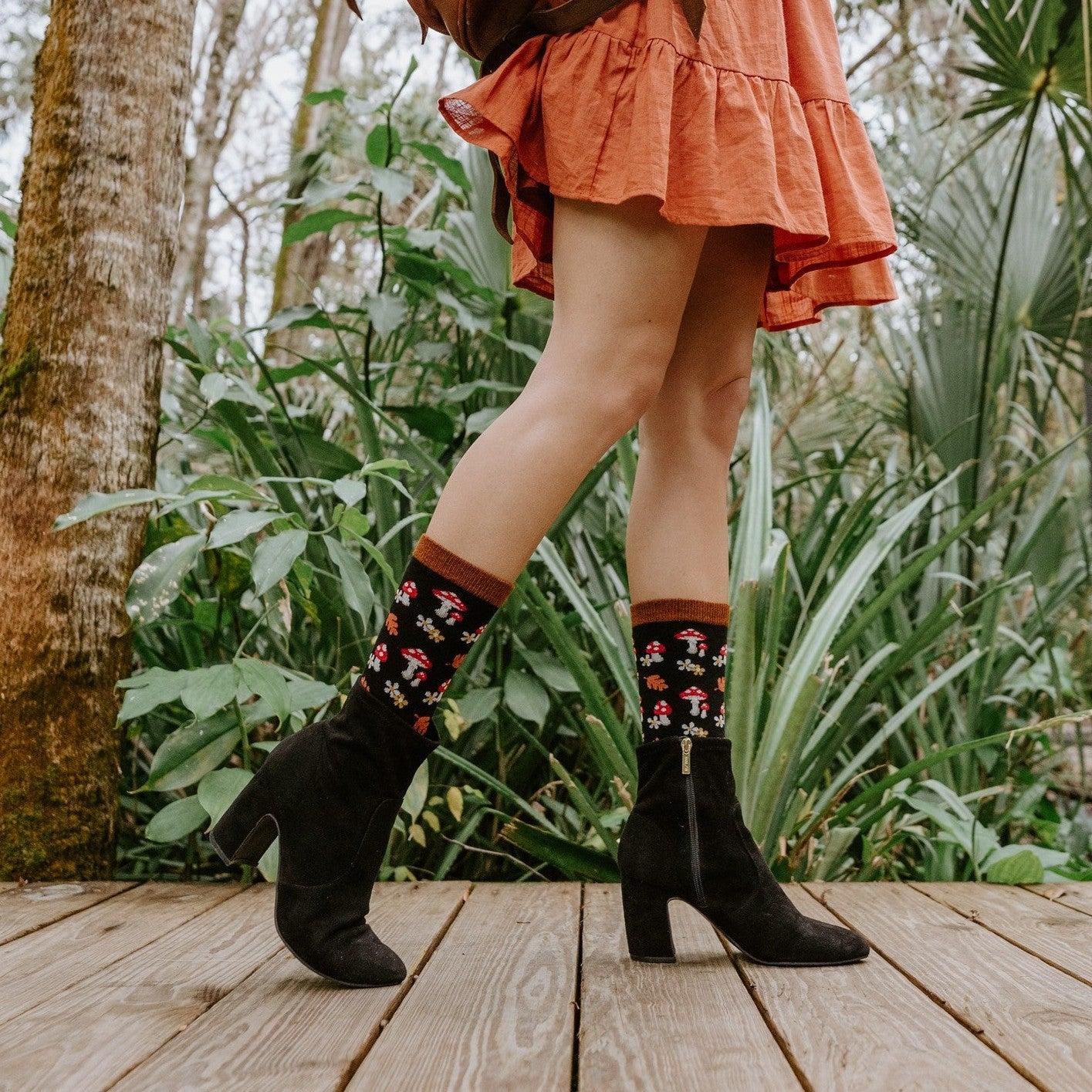 A pair of black heeled booties and Gems Of The Forest Bamboo socks in Black on a wood deck, the pattern of mushrooms and daisies clearly visible at the cuff. [lf]