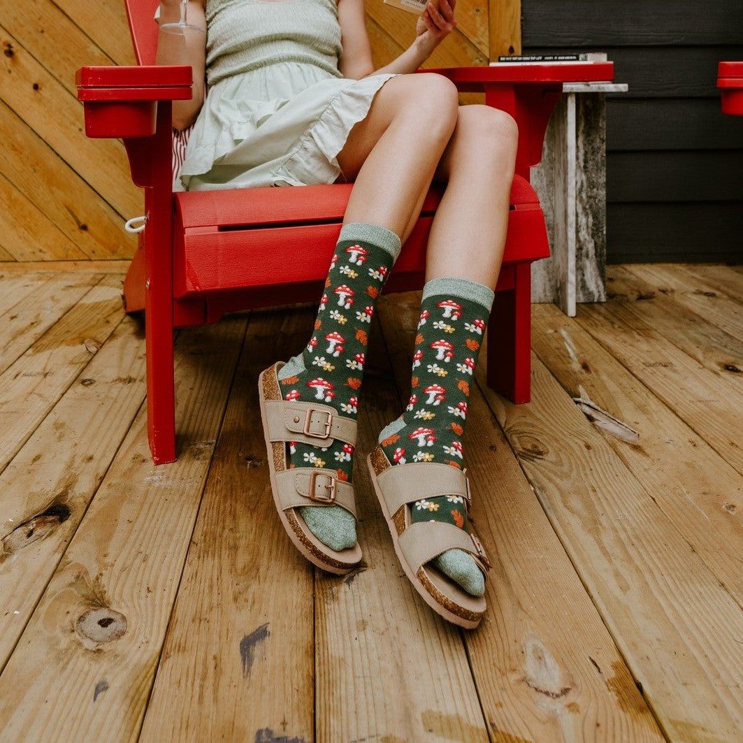 A woman in a sage dress reads a book on a sunlit porch, her crossed legs showing Gems Of The Forest Bamboo socks in Black with mushroom and floral details. [lf]