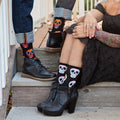 A close-up of Dia De Los Muertos Skull socks in White on a person seated by an altar decorated with painted skulls. [lf]
