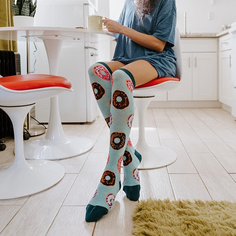 A pair of Go Nuts For Donuts socks in Blue Heather crossed on a rug beside a pink donut box. [lf]