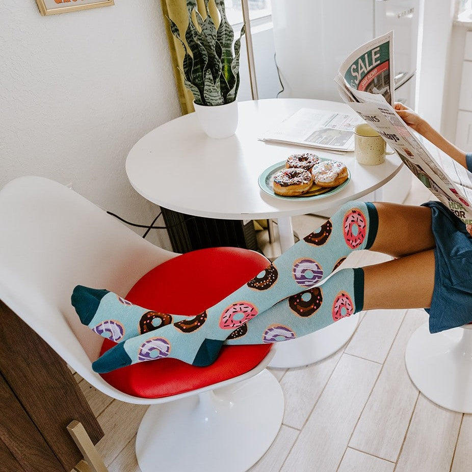 A friend laughs holding up a donut, her Go Nuts For Donuts socks in Blue Heather kicked out across the cafe floor. [lf]