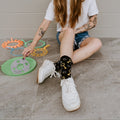 A close-up of Blowing Bubbles socks in Black featuring bubble illustrations, on a person seated on a sunny porch with a bubble wand. [lf]