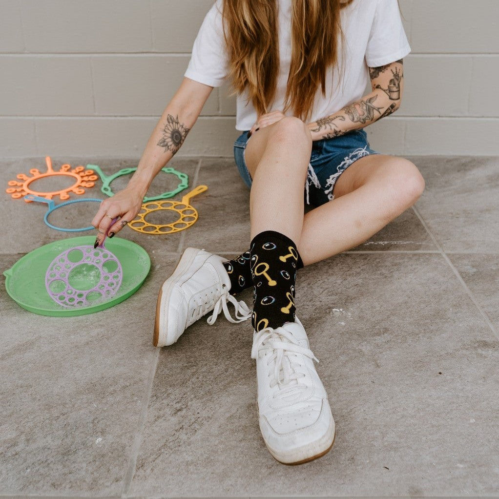 A close-up of Blowing Bubbles socks in Black featuring bubble illustrations, on a person seated on a sunny porch with a bubble wand. [lf]