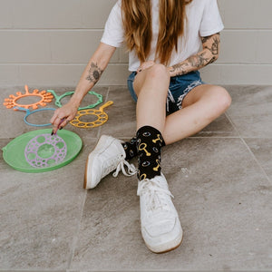A close-up of Blowing Bubbles socks in Black featuring bubble illustrations, on a person seated on a sunny porch with a bubble wand. [lf]