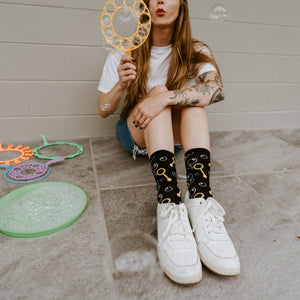 A close-up of Blowing Bubbles socks in Black on a person dipping a bubble wand into solution. [lf]