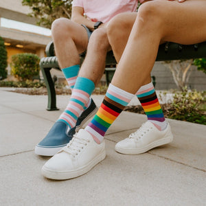 A close-up of Progress Stripes socks in Multi on a wood porch with a rainbow flag fluttering in the background. [lf]