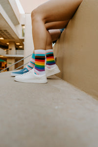 A close-up of Progress Stripes socks in Multi resting on a yoga mat after a workout, paired with athletic sneakers. [lf]