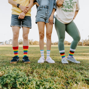A close-up of Trans Stripes socks in Multi featuring the trans-pride color stripes, on a person crossing a sunny sidewalk. [lf]