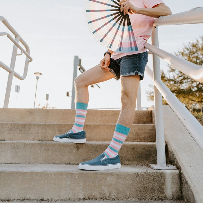 A pair of Trans Stripes socks in Multi stretched on a grassy lawn beside a small flag. [lf]