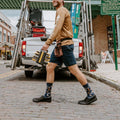 A pair of Can You Fix It? socks in Navy Heather on a man's crossed feet propped on a workshop stool, surrounded by hand tools. [lf]