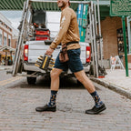 A pair of Can You Fix It? socks in Navy Heather on a man's crossed feet propped on a workshop stool, surrounded by hand tools. [lf]
