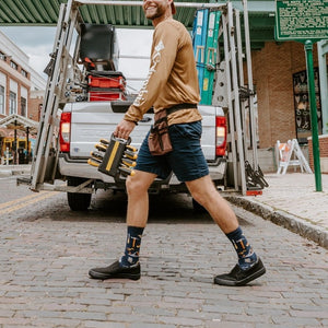 A pair of Can You Fix It? socks in Navy Heather on a man's crossed feet propped on a workshop stool, surrounded by hand tools. [lf]