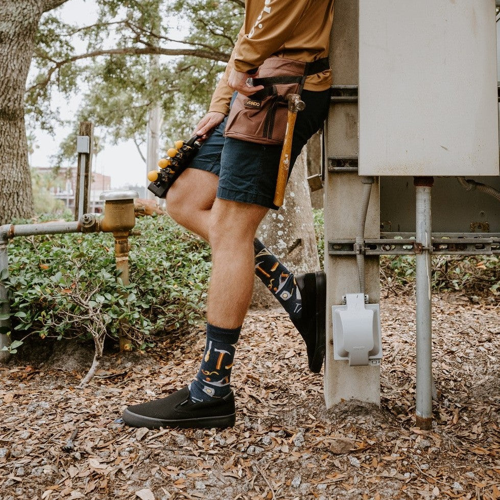 A friend hands a tool across a workbench while modeling Can You Fix It? socks in Navy Heather with hammer and saw graphics. [lf]