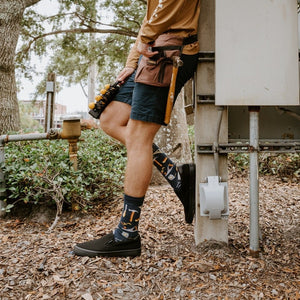 A friend hands a tool across a workbench while modeling Can You Fix It? socks in Navy Heather with hammer and saw graphics. [lf]