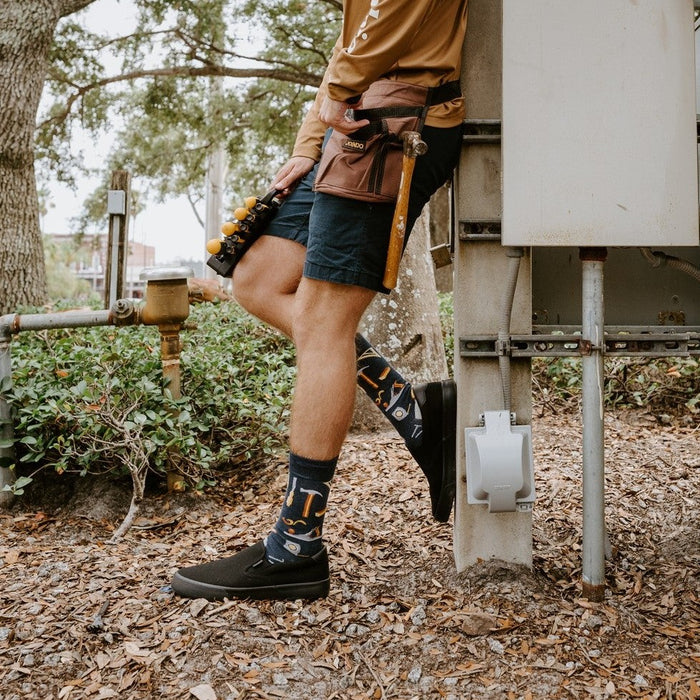 A friend hands a tool across a workbench while modeling Can You Fix It? socks in Navy Heather with hammer and saw graphics. [lf]
