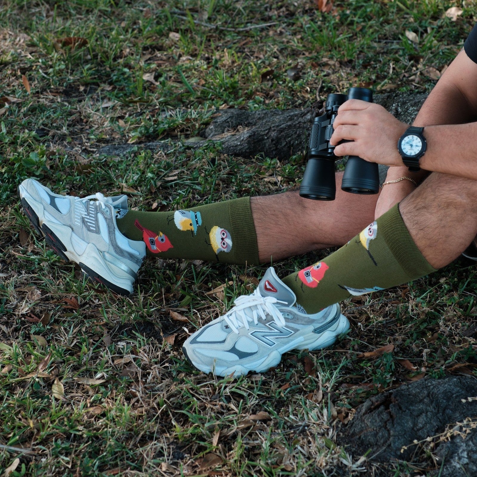 A pair of Bird Is The Nerd socks in Green crossed on a chair beside a stack of books and a bird feeder. [lf]