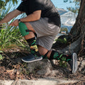 A friend in a colorful outfit poses against a mural, modeling Bright As A Chameleon socks in Black with chameleon prints. [lf]