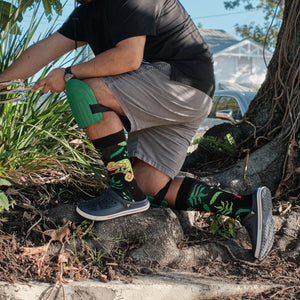 A friend in a colorful outfit poses against a mural, modeling Bright As A Chameleon socks in Black with chameleon prints. [lf]