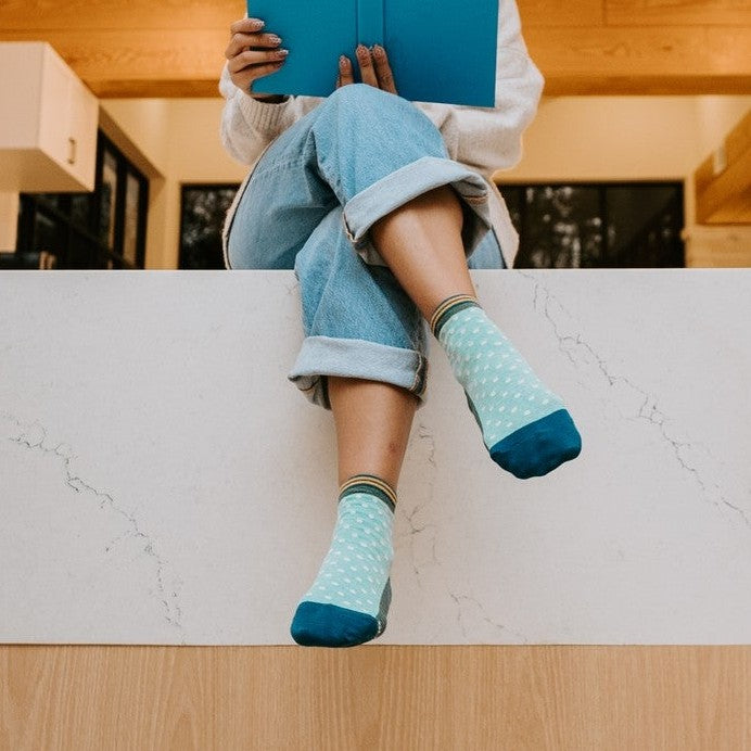 A close-up of Pin Dot socks in Teal Heather featuring tiny pin-dot patterns, paired with loafers on a polished wood floor. [lf]