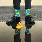 A model stands by a puddle in cuffed grey jeans, wearing Rubber Ducky blue socks with yellow rubber duck prints above polished black Oxford shoes beside a real yellow rubber duck. [lf]