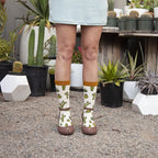 A close-up of crossed feet in King Cactus Bamboo cream socks with cactus and crown prints, posed against a backdrop of potted cacti and succulents. [lf]
