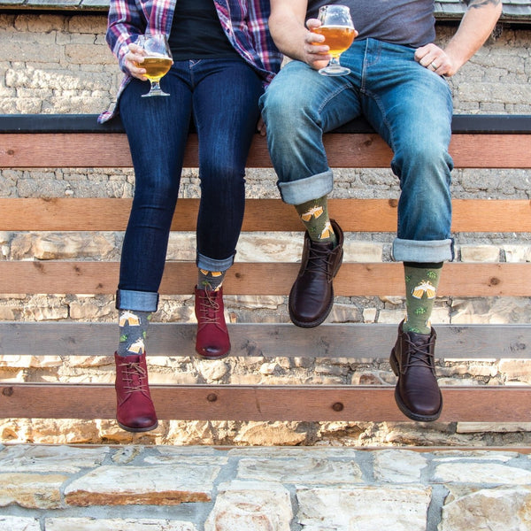 A seated couple share craft beers on a wooden plank bench in Hoppier Together socks featuring beer mug prints, paired with red and brown leather boots. [lf]
