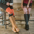A woman sips coffee on a metal stool wearing Fox orange knee-high socks featuring a smiling fox face, tucked into tan riding boots beside a friend in pink cat-face socks. [lf]