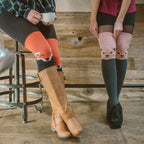 A woman sips coffee on a metal stool wearing Fox orange knee-high socks featuring a smiling fox face, tucked into tan riding boots beside a friend in pink cat-face socks. [lf]