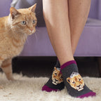 A woman sits on a purple couch with an orange tabby cat at her side, wearing Queen of the Pride grey ankle socks with regal lioness portraits crowned in lace. [lf]