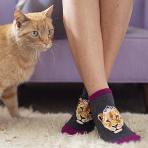 A woman sits on a purple couch with an orange tabby cat at her side, wearing Queen of the Pride grey ankle socks with regal lioness portraits crowned in lace. [lf]