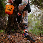 A tattooed model with a jack-o-lantern mask sits cross-legged on a black blanket, wearing Halloween Icons black socks with orange ghost and candy prints. [lf]