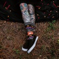 Halloween Icons socks shown on a tattooed model posed playfully in a forest with carved pumpkins. [lf]