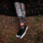 Halloween Icons socks shown on a tattooed model posed playfully in a forest with carved pumpkins. [lf]
