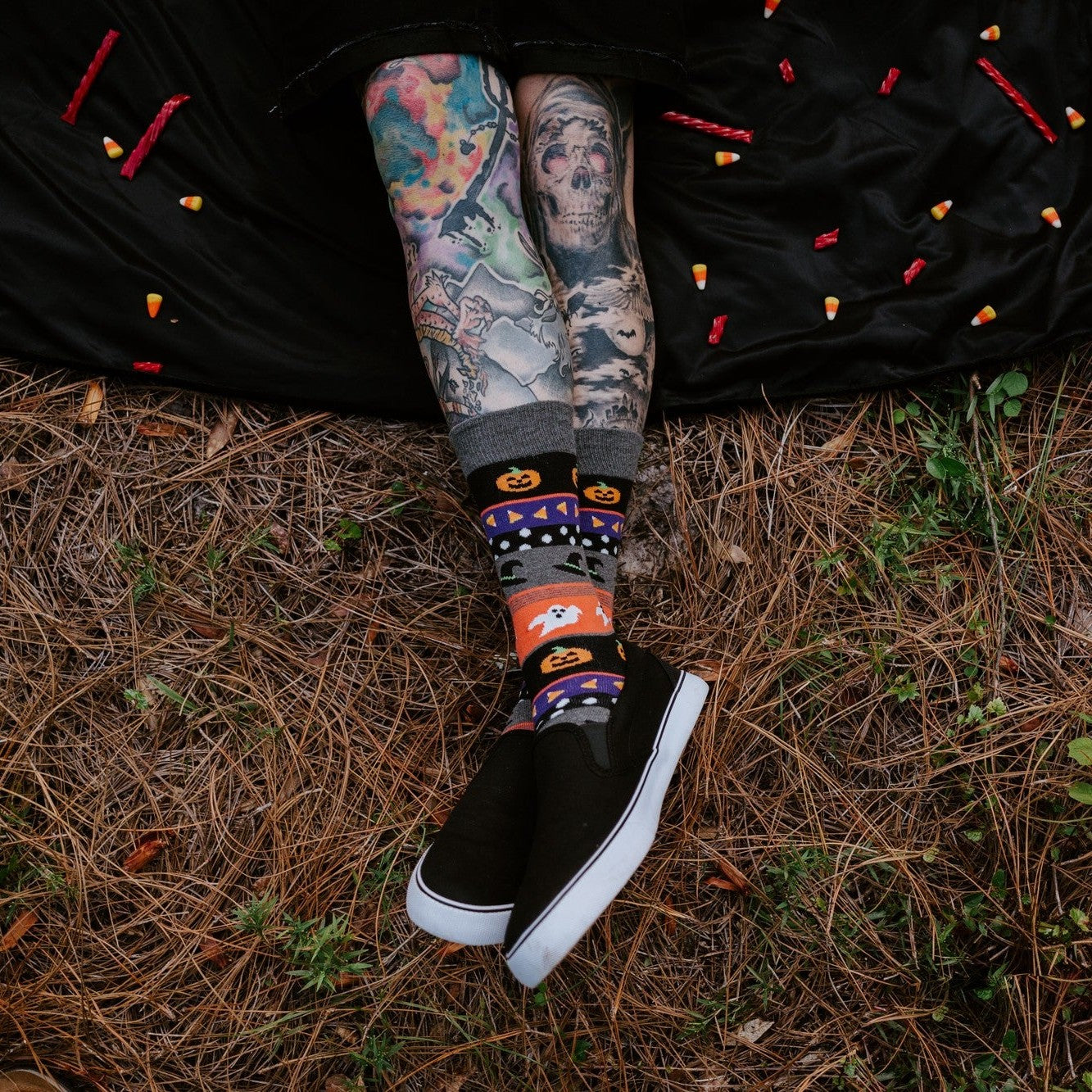 Halloween Icons socks shown on a tattooed model posed playfully in a forest with carved pumpkins. [lf]