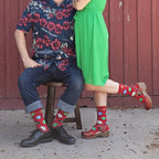 A couple poses against a red barn door in coordinating Christmas outfits, both wearing Ugly Sweaters red holiday socks with poinsettia and ornament motifs. [lf]