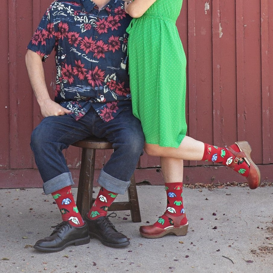 A couple poses against a red barn door in coordinating Christmas outfits, both wearing Ugly Sweaters red holiday socks with poinsettia and ornament motifs. [lf]
