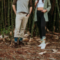 A couple stands among tall bamboo, the man in olive top and chinos wearing Honey In The Bank Bamboo socks with bees and sunflowers, paired with brown boots. [lf]