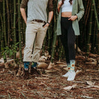 A couple stands among tall bamboo, the man in olive top and chinos wearing Honey In The Bank Bamboo socks with bees and sunflowers, paired with brown boots. [lf]