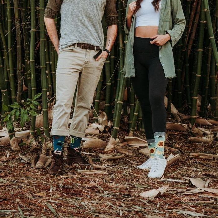 A couple stands among tall bamboo, the man in olive top and chinos wearing Honey In The Bank Bamboo socks with bees and sunflowers, paired with brown boots. [lf]