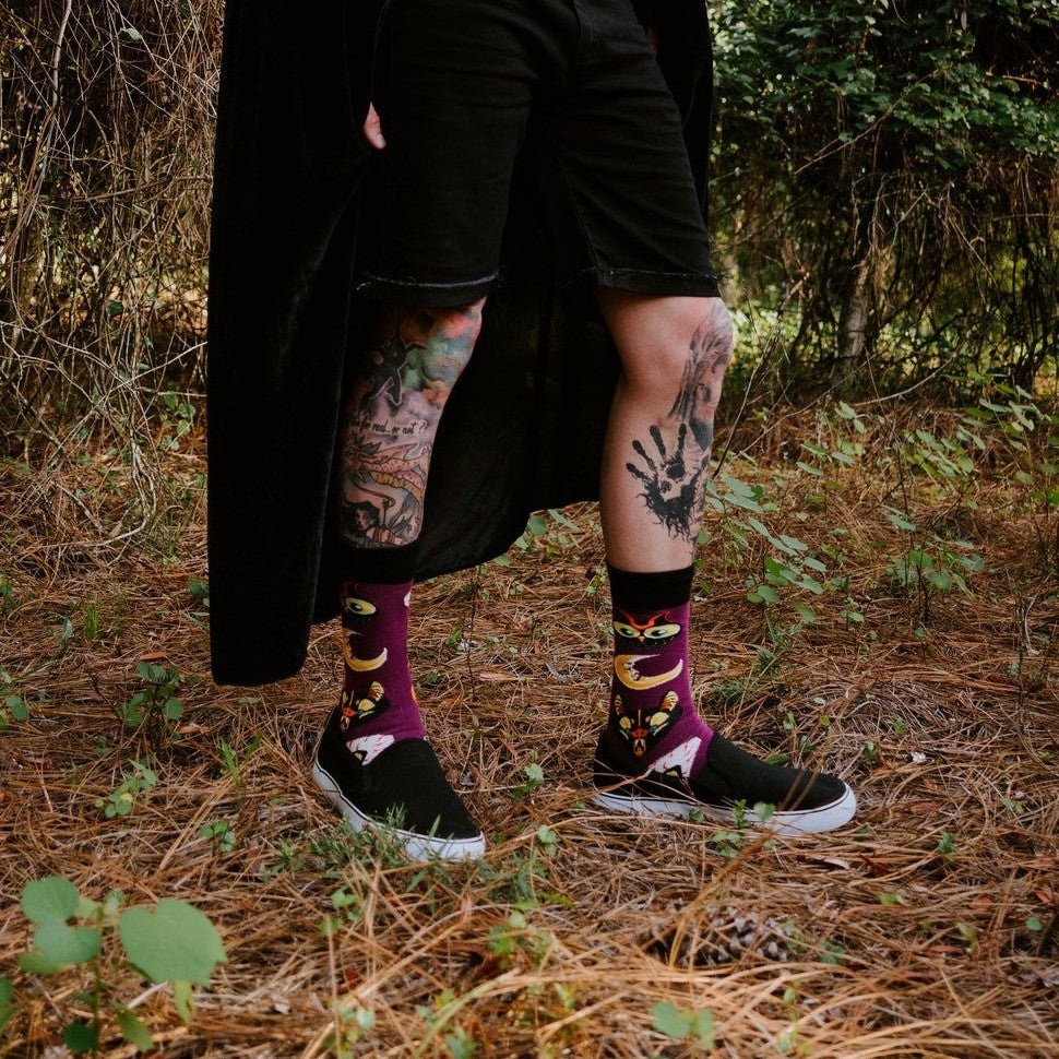 All Hallows Eve socks displayed on a model crouched beside a cauldron, surrounded by candles and a fake skull. [lf]