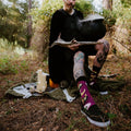 All Hallows Eve socks worn by a tattooed model staging a witchy outdoor scene with candles, cauldron, and skull. [lf]