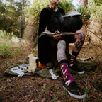 All Hallows Eve socks worn by a tattooed model staging a witchy outdoor scene with candles, cauldron, and skull. [lf]