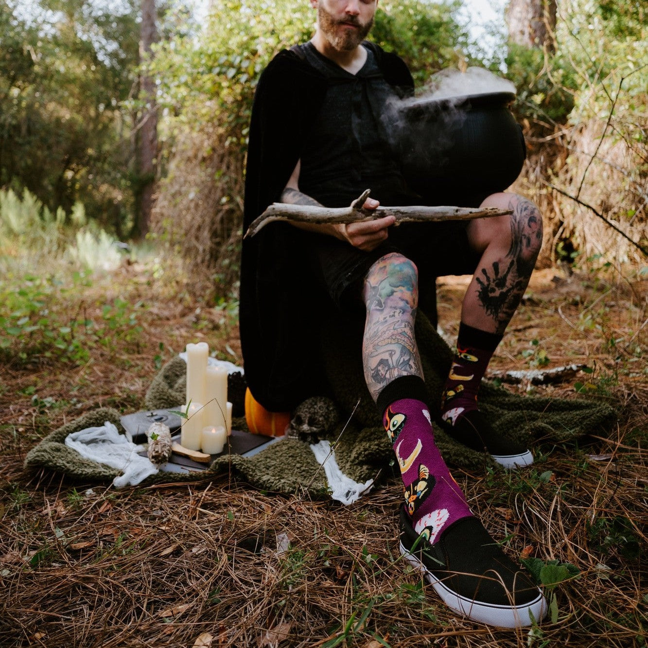 All Hallows Eve socks worn by a tattooed model staging a witchy outdoor scene with candles, cauldron, and skull. [lf]