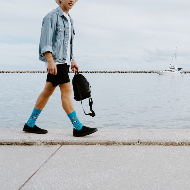A man in a denim jacket and straw hat walks along a marina pier wearing Don't Rock The Boat blue nautical socks above black slip-on shoes. [lf]