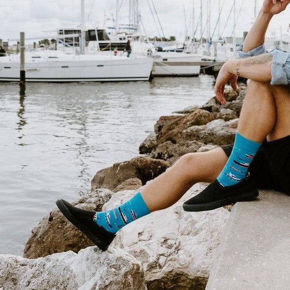 A close-up of Don't Rock The Boat blue socks with sailboat prints worn with black canvas shoes on a sunny waterfront walk. [lf]