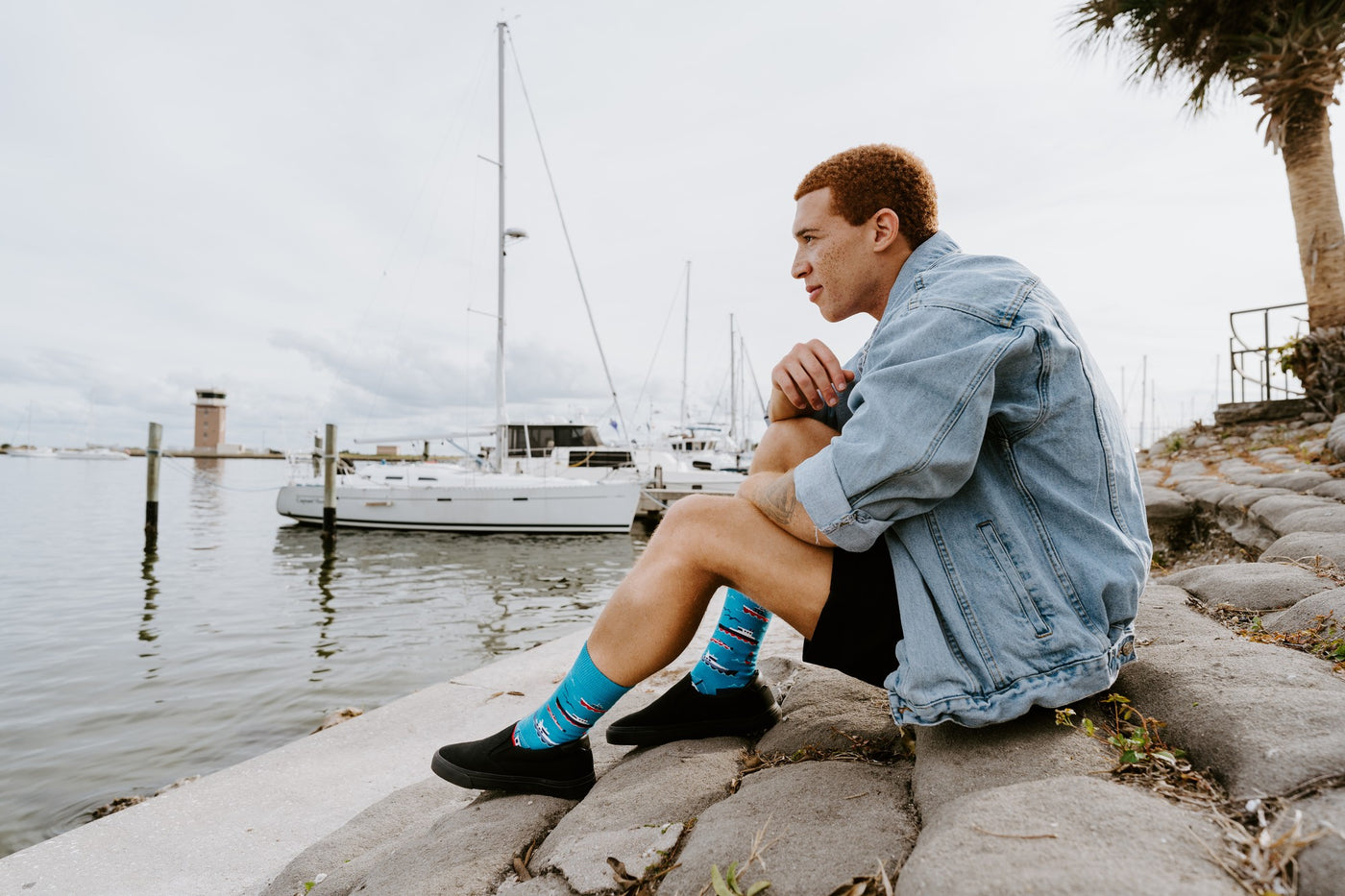 A man in a denim jacket and straw hat walks along a marina pier wearing Don't Rock The Boat blue nautical socks above black slip-on shoes. [lf]